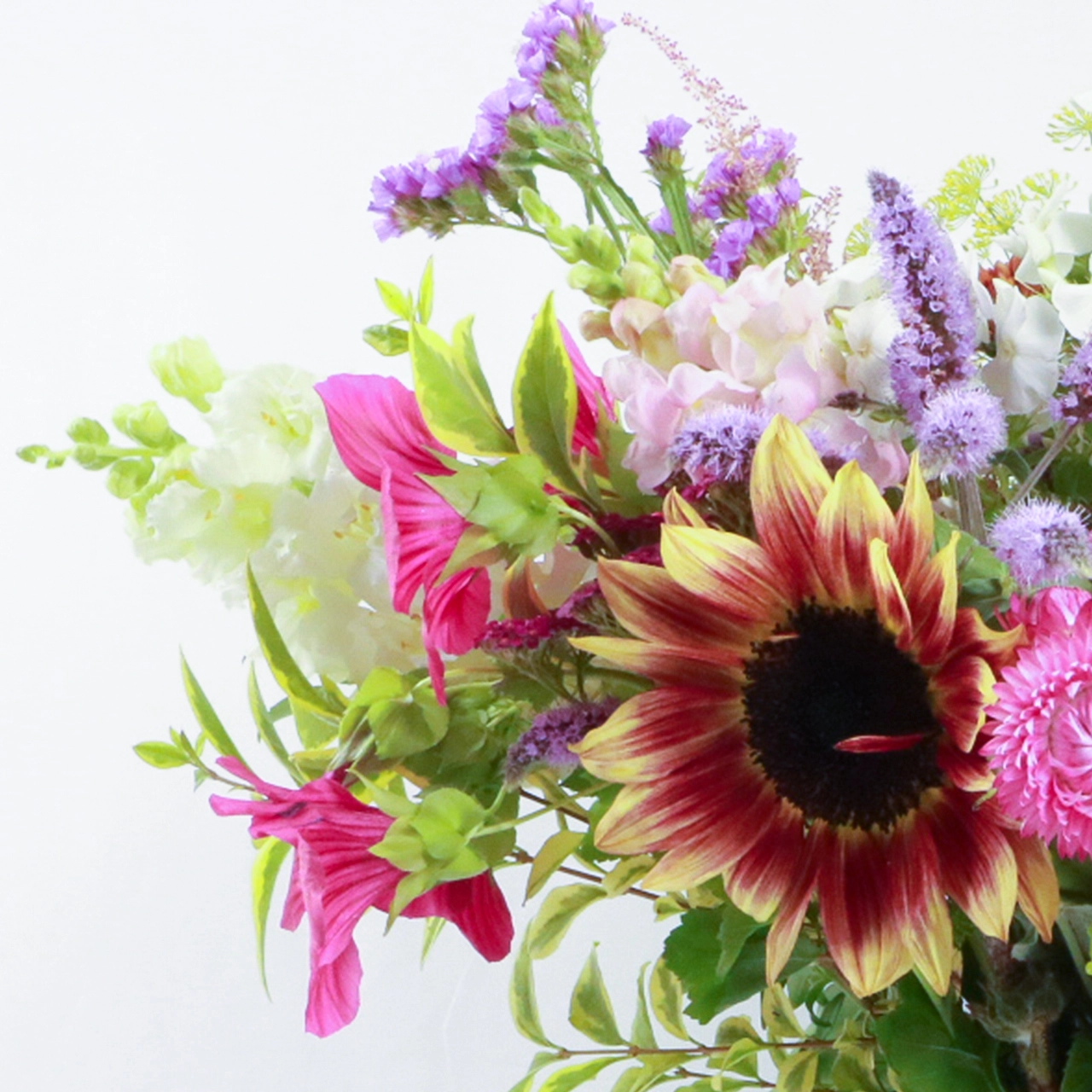 Close-up detail of Marguerite Rose Devonshire Deluxe bouquet showing red-yellow sunflower, pink cosmos, purple wildflowers and seasonal foliage