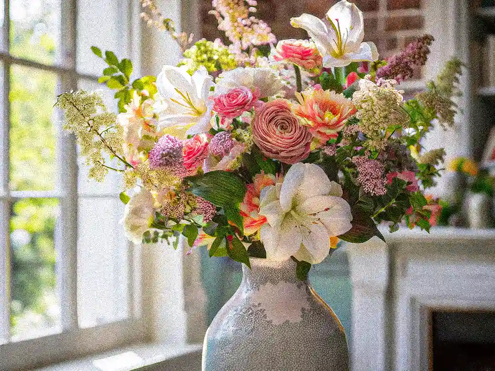 A stunning, vibrant mixed flower bouquet with lilies, roses, and various blooms beautifully arranged in a vase by a window, ready for enjoyment from Marguerite Rose.