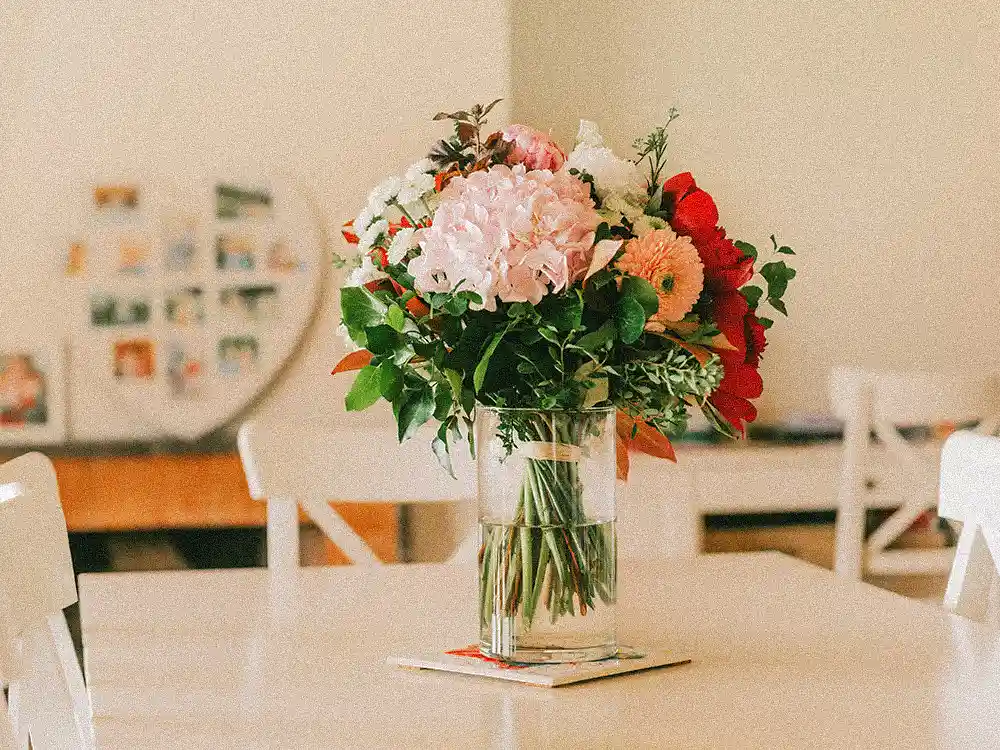 A vibrant mixed flower bouquet in a clear vase sits on a light table, demonstrating ideal placement away from direct sun for Marguerite Rose flowers.