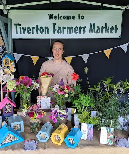 Marguerite Rose stands smiling behind her stall at Tiverton Farmers Market, surrounded by colorful bouquets of flowers, potted plants, and various styles of bee hotels. A sign above her reads "Welcome to Tiverton Farmers Market".